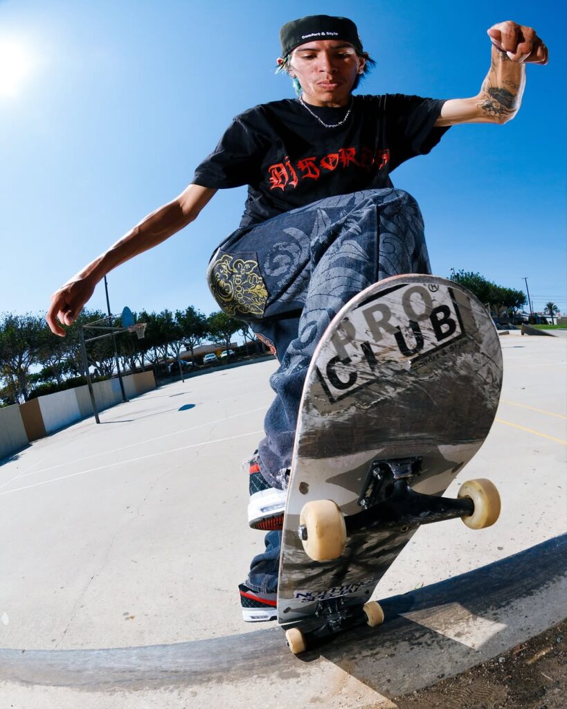 Un skater mexicano realiza un truco de calle sobre un borde urbano, vistiendo ropa holgada de estilo Pro Club y sosteniendo una tabla Nothing Special frente a una cancha local.