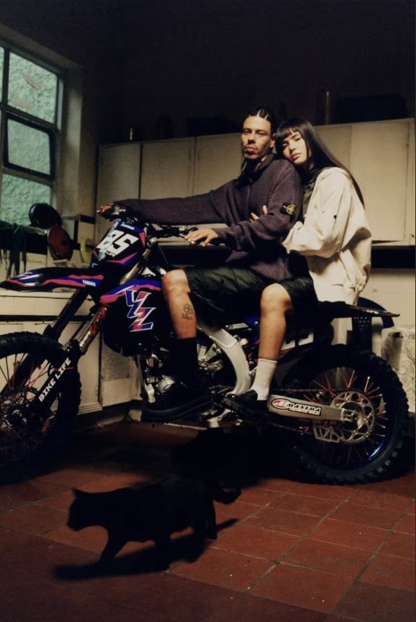 Una pareja posando sobre una motocicleta de motocross Yamaha azul con el texto Bike Life escrito en la suspensión delantera, vistiendo suéteres Stone Island dentro de una cocina rústica en Medellín, Colombia, con un gato negro caminando en primer plano.