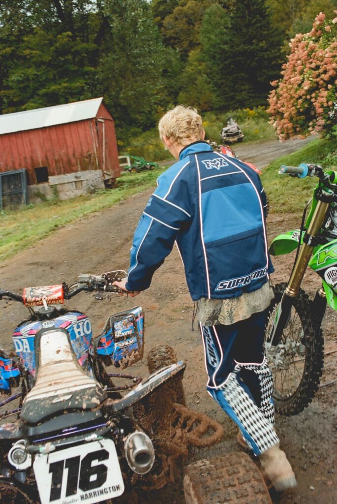 Joven rubio de espaldas con chaqueta azul de carreras Supreme y Fox Racing caminando entre un cuatrimoto Honda cubierto de lodo con el número 116 y una motocicleta de motocross Kawasaki verde.