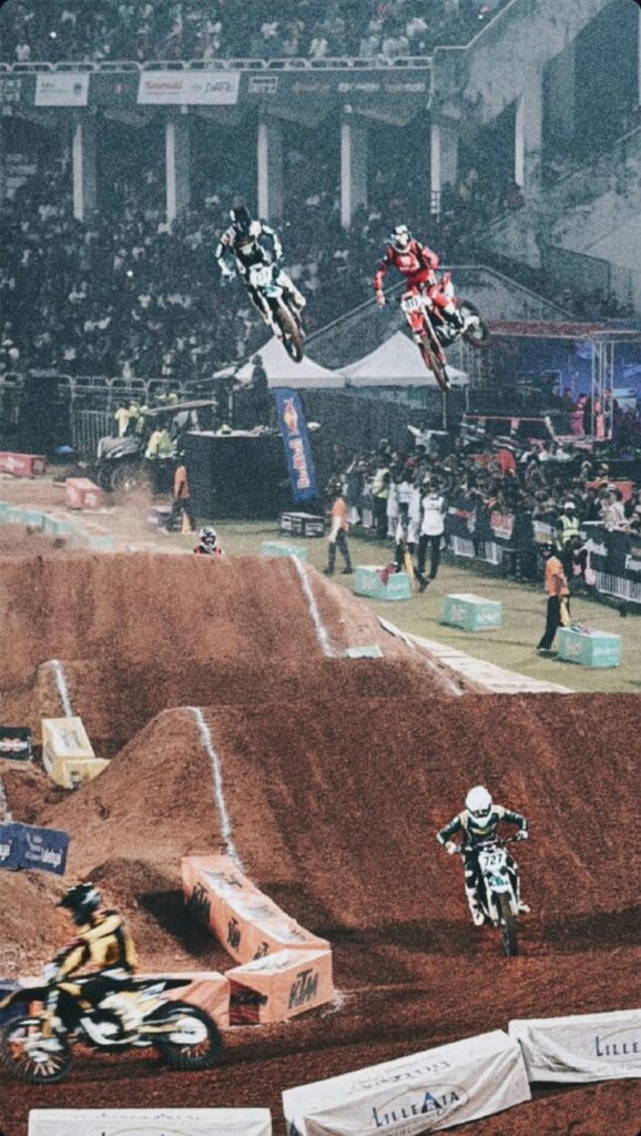 Pilotos profesionales de la Indian Supercross Racing League volando en paralelo sobre rampas de tierra roja frente a las gradas de un estadio lleno durante una competencia nocturna en India.