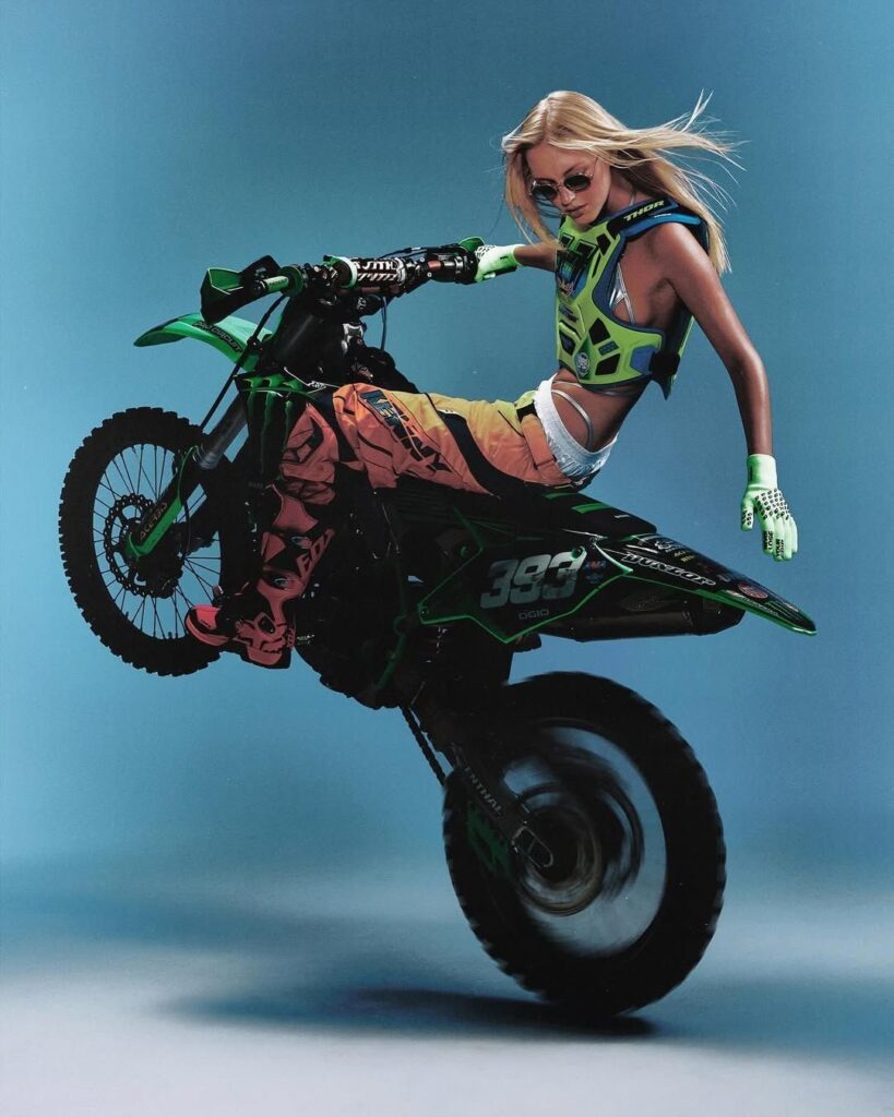 Modelo rubia posando con gafas de sol, guantes de carreras verdes y chaleco protector Thor MX inclinada sobre el asiento de una motocicleta de motocross Kawasaki verde en movimiento.