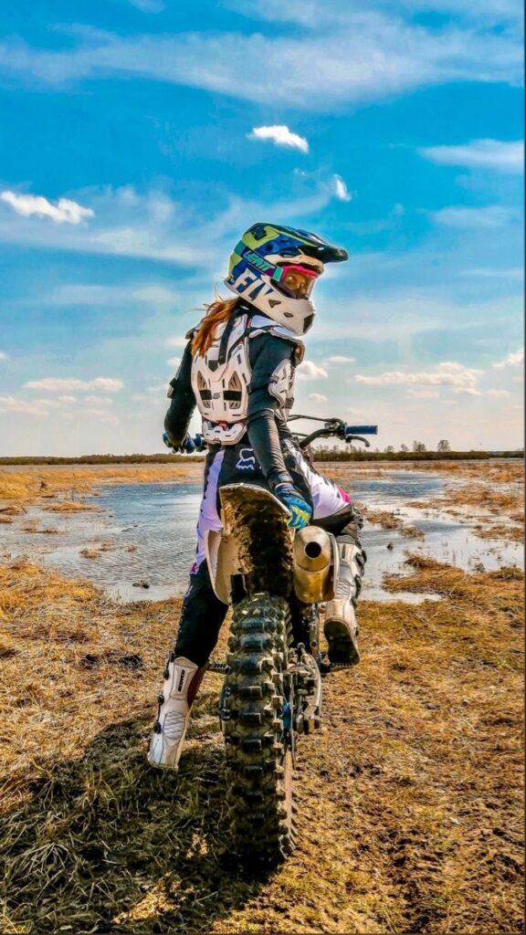 Vista trasera de una piloto de motocross posando de pie sobre una motocicleta de cross en un camino de tierra húmeda y lodo bajo un cielo azul con nubes.
