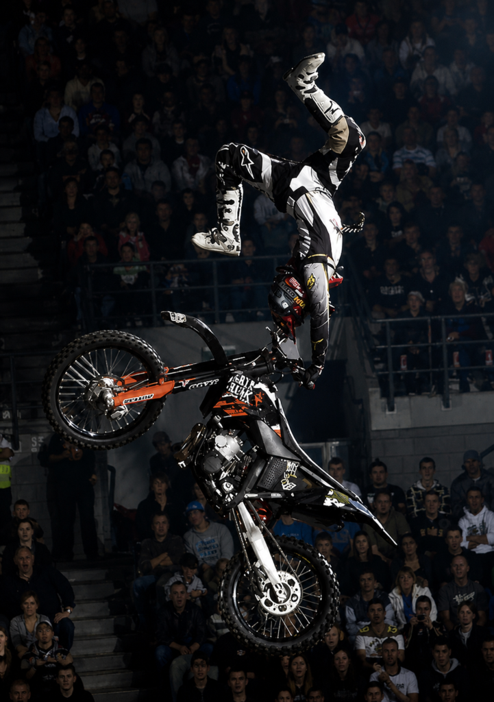 Piloto de Freestyle Motocross realizando un truco invertido colgado del manubrio de su motocicleta KTM negra durante la competencia internacional Night of the Jumps en un estadio cubierto lleno de público.