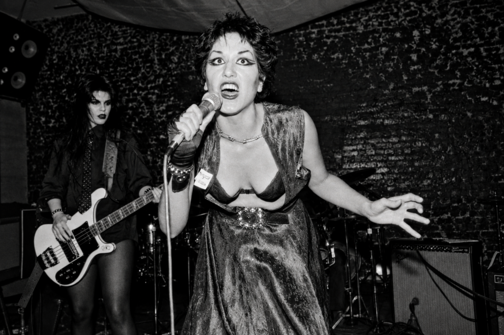 Alice Bag cantando intensamente al micrófono junto a la bajista Patricia Morrison durante un concierto en vivo de la banda punk The Bags en un club underground de Los Ángeles en 1978.