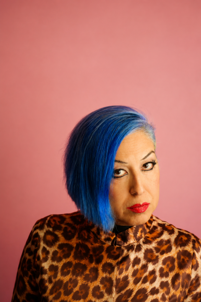 Retrato moderno de estudio de la legendaria cantante de punk chicano Alice Bag, posando con su icónico cabello teñido de azul brillante, labios rojos y una chaqueta con estampado de leopardo sobre un fondo rosa pastel.