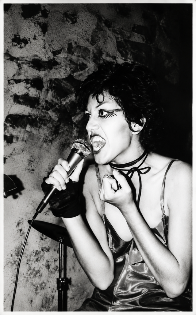 Retrato en blanco y negro de la cantante chicana Alice Bag gritando con furia punk al micrófono, luciendo su icónico delineado de ojos egipcio y un lazo negro al cuello durante un concierto en Los Ángeles en 1978.