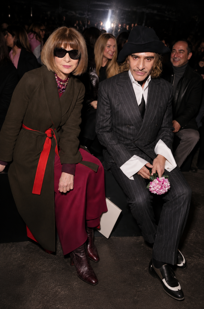 Anna Wintour con gafas de sol oscuras y abrigo verde oliva junto al diseñador John Galliano vistiendo un traje de raya diplomática y sombrero negro, sentados en la primera fila de un desfile de modas en París.