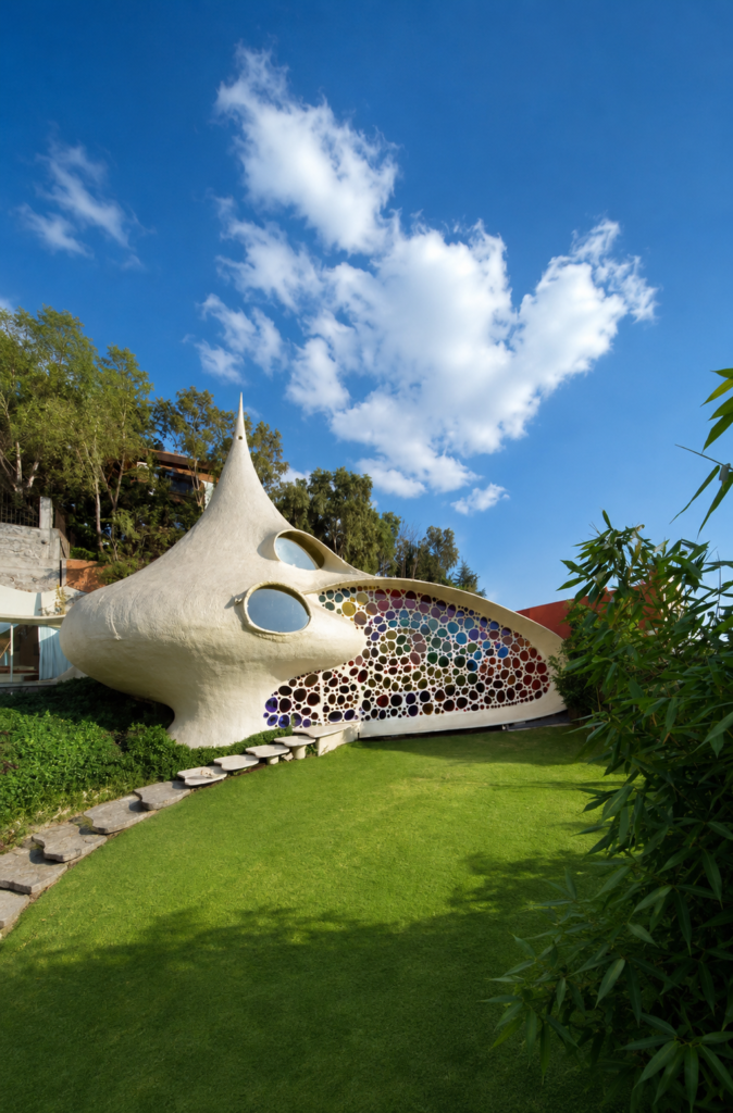 Fachada exterior de la Casa Nautilus en Naucalpan, México, obra de arquitectura orgánica diseñada por Javier Senosiain con forma de caracol y un gran vitral de mosaicos coloridos en un jardín verde bajo el cielo azul.