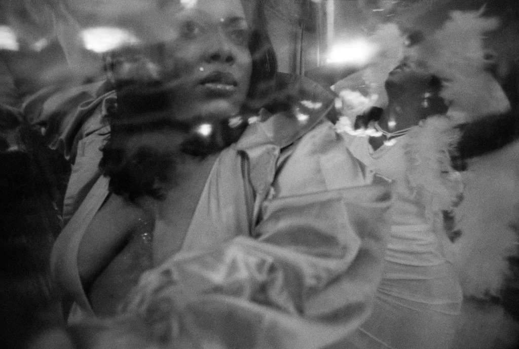 Fotografía artística en blanco y negro que muestra el reflejo en un espejo distorsionado de una mujer afroamericana con un vestido de satén escotado mirando fijamente hacia un lado. Al fondo, a través del reflejo, se aprecia a otra persona bailando con una boa de plumas blancas.