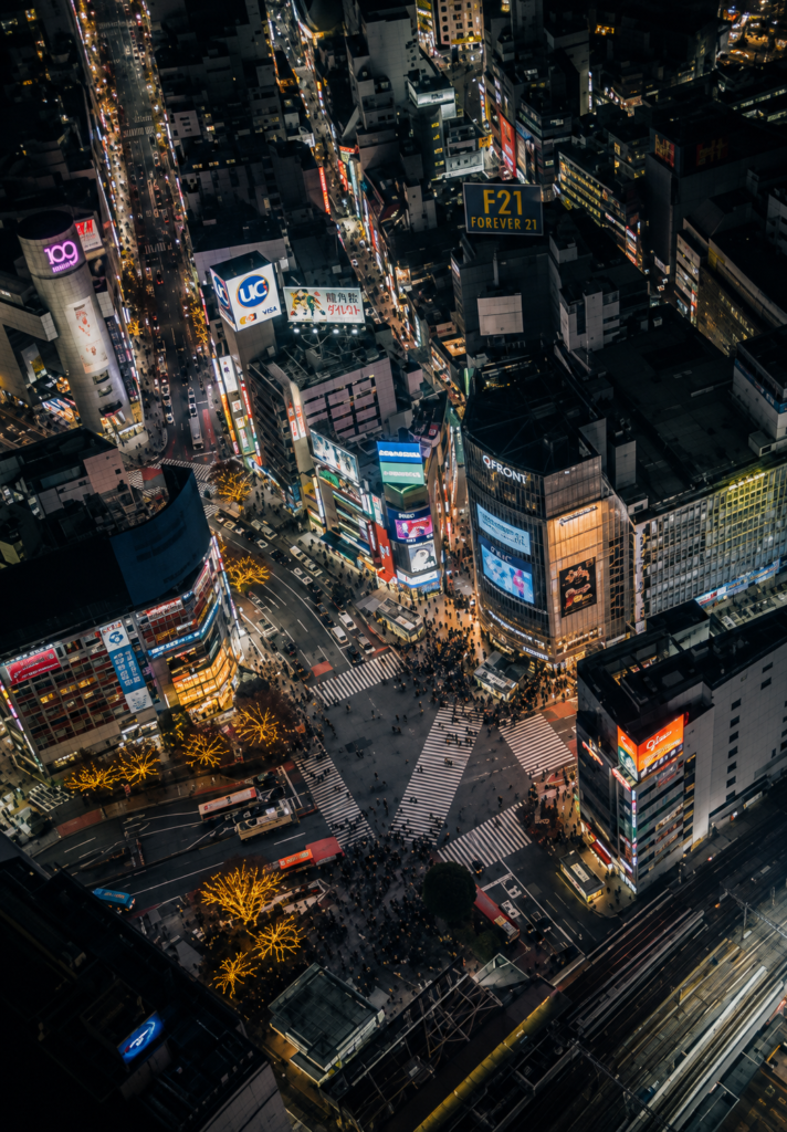 Vista aérea nocturna del icónico cruce de Shibuya en Tokio repleto de peatones, luces de neón y pantallas gigantes de edificios como el QFRONT, capturando la esencia futurista y cyberpunk ideal para rutas de turismo literario inspiradas en William Gibson.