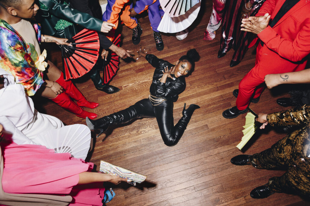 Vista aérea de una mujer afroamericana sonriendo en el suelo mientras realiza una pose de voguing en la categoría de pasarela. Viste un traje completo de cuero negro ajustado con botas de tacón altas, rodeada por un círculo de personas y espectadores que aplauden sosteniendo abanicos plegables rojos y negros sobre un suelo de madera.