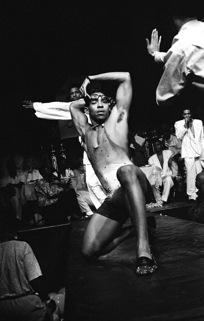 Fotografía en blanco y negro de un bailarín afroamericano haciendo una pose de voguing en una pasarela durante un ball en Nueva York. Lleva lentes oscuros y un atuendo entallado con pedrería, rodeado de público y jueces al fondo.