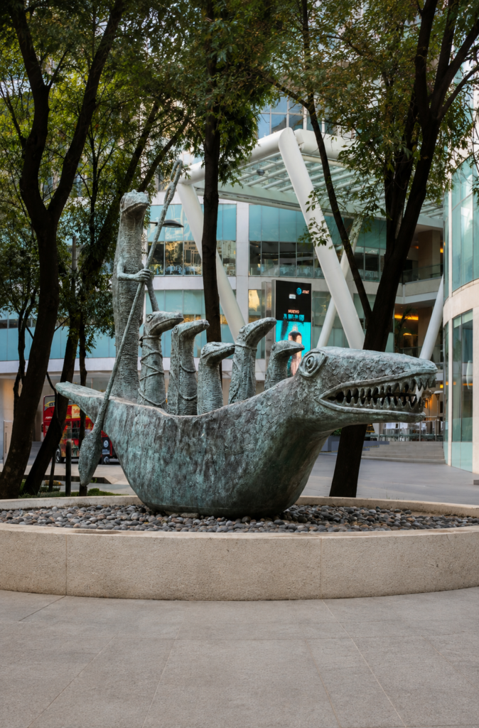 Estatua de bronce "El Cocodrilo" de Leonora Carrington en Paseo de la Reforma, Ciudad de México, mostrando una embarcación con forma de cocodrilo gigante y pequeñas criaturas a bordo en medio de una plaza urbana.