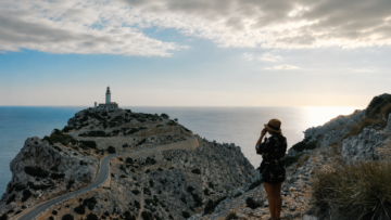 Turismo literario: rutas Beat, noir y distópicas para viajar siguiendo a escritores de culto Una turista con sombrero observa el Faro de Cap de Formentor en un acantilado rocoso de Mallorca, España, un destino histórico de turismo literario.
