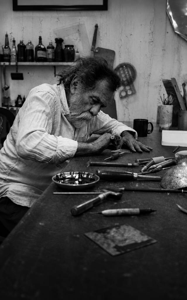 Fotografía vertical en blanco y negro del maestro Francisco Toledo concentrado tallando una placa de grabado con gubias en la mesa de su taller.