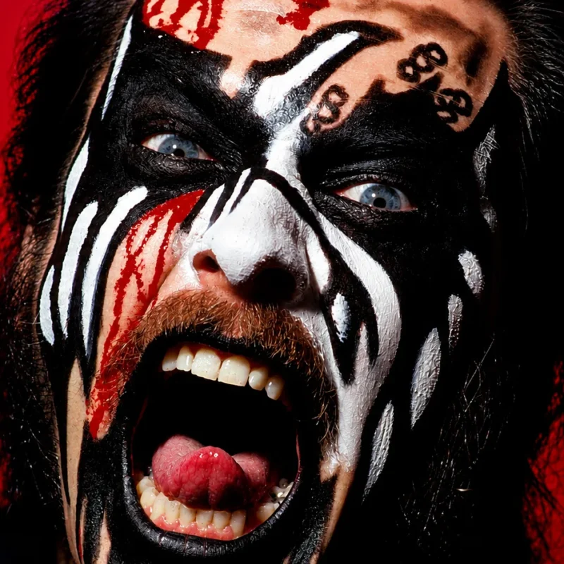 Primer plano de King Diamond con su maquillaje de corpse paint característico, incluyendo detalles de sangre simulada, gritando furiosamente a la cámara sobre un fondo rojo intenso.