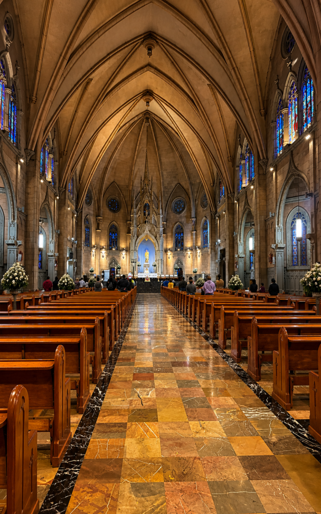 Interior del Santuario Nacional de María Auxiliadora en CDMX, con simétricas bóvedas de crucería neogóticas, bancas de madera alineadas, piso de mármol y vitrales de colores.