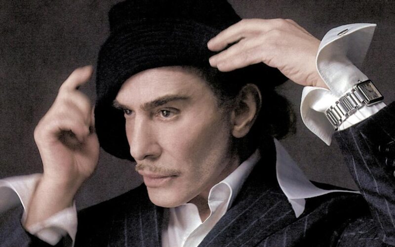 Retrato de perfil del diseñador de moda John Galliano con sombrero negro, traje de raya diplomática y reloj de pulsera plateado, fotografiado por Szilveszter Makó.