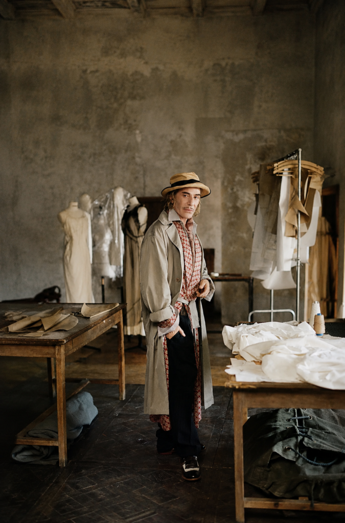 Diseñador John Galliano de cuerpo entero con gabardina beige y sombrero en su taller de costura rodeado de maniquíes y patrones de ropa en mesas de madera.