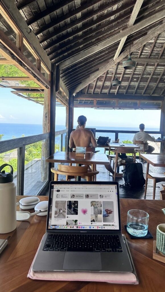 Perspectiva en primera persona de un espacio de coworking en una cafetería tropical frente al mar, con una computadora portátil MacBook abierta mostrando la plataforma de inspiración Pinterest en la pantalla. Sobre la mesa de madera hay un termo de agua, auriculares inalámbricos blancos y un vaso de agua; al fondo se observan otros nómadas digitales trabajando en un entorno abierto con vistas al océano.