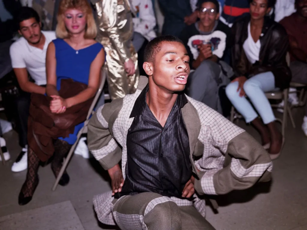 Primer plano de un joven afroamericano bailando activamente voguing en un salón. Viste una camisa negra semiabierta y un abrigo de cuadros beige y verde con las manos apoyadas en su cadera. Al fondo, varios espectadores sentados lo observan atentos, incluyendo a una mujer rubia con un vestido azul.