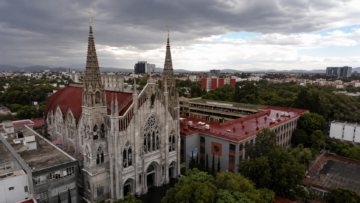 Guía práctica para explorar la arquitectura neogótica en la Ciudad de México Vista aérea con dron del Santuario Nacional de María Auxiliadora en CDMX, destacando sus dos altas torres caladas, fachada de cantera gris y techos rojos de estilo neogótico italiano.