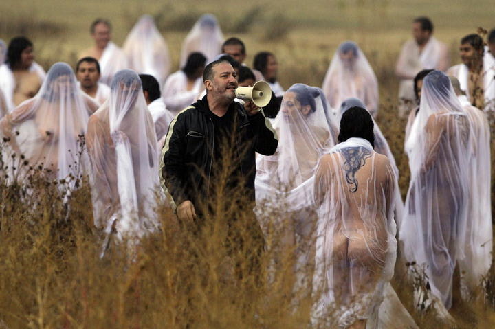 El fotógrafo Spencer Tunick con un megáfono dirigiendo a personas desnudas cubiertas individualmente por velos y bolsas transparentes en un campo abierto, como parte de su instalación artística "Inversion" adaptada a las medidas de distanciamiento físico por la pandemia.