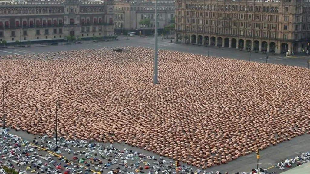 Toma elevada de miles de personas desnudas en posición fetal en la Plaza de la Constitución o Zócalo de la Ciudad de México, con montones de ropa amontonada en la esquina inferior izquierda, instalación masiva de Spencer Tunick.