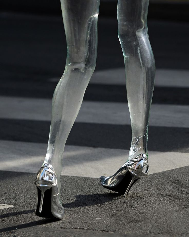 Primer plano detallado de piernas esculpidas en cristal transparente simulando caminar sobre un paso peatonal, calzando zapatos de tacón aguja cerrados y metalizados en cromo plateado.