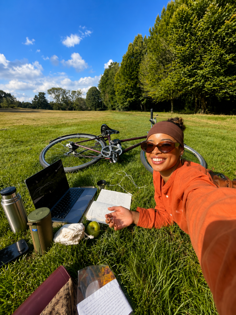 Una mujer joven sonriente tomándose una selfie mientras está recostada en el césped de un parque verde en un día soleado. A su lado tiene una computadora portátil abierta, un cuaderno de notas, un termo de agua, una manzana verde y una bicicleta de montaña recostada en el pasto, ilustrando el concepto de teletrabajo al aire libre o nómada digital.