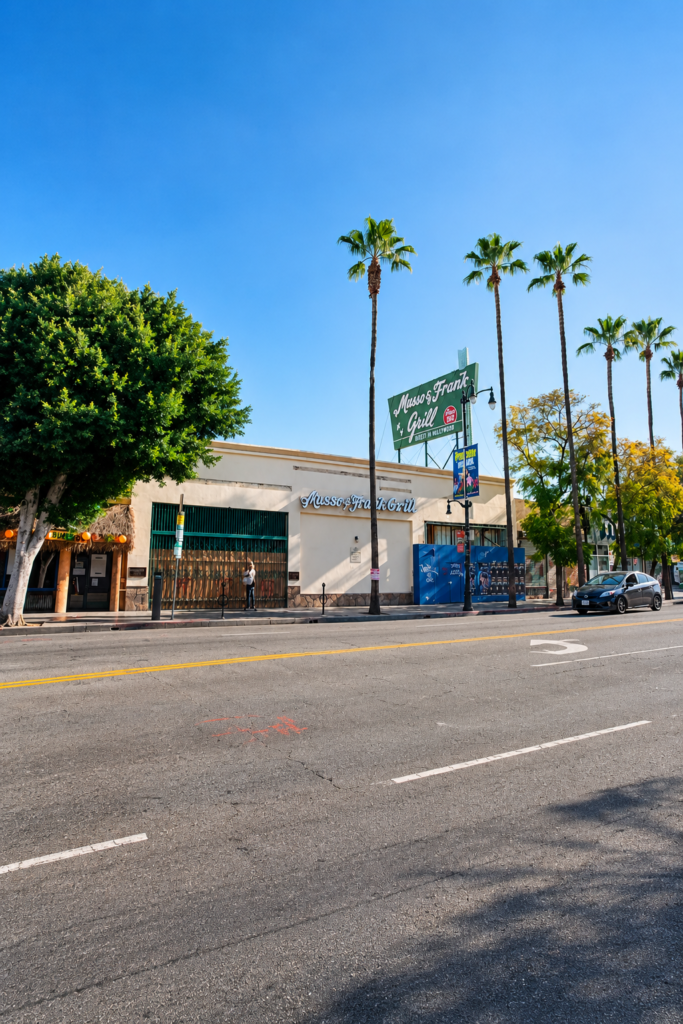 Fachada exterior del restaurante histórico Musso and Frank Grill en Hollywood Boulevard, Los Ángeles, Estados Unidos, mostrando su icónico letrero verde entre palmeras altas bajo un cielo despejado.