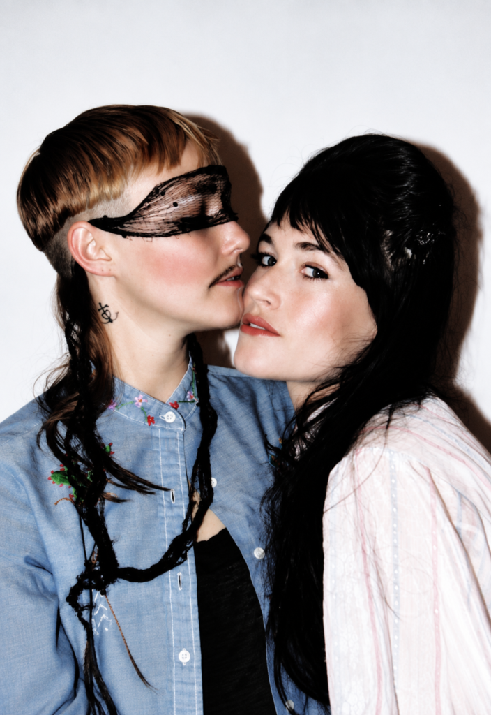 Bianca y Sierra Casady de CocoRosie posan juntas con pelucas negras y una máscara de encaje en un retrato promocional.