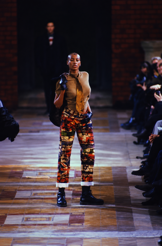 Look de Jean Paul Gaultier de 1994 con pantalones estampados, top utilitario, corbata, guantes y botas negras en pasarela.