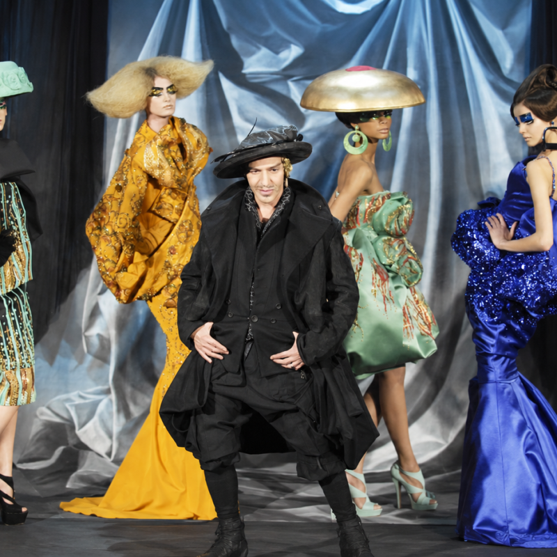John Galliano posa rodeado de modelos con vestidos escultóricos y sombreros teatrales durante un desfile de alta costura.