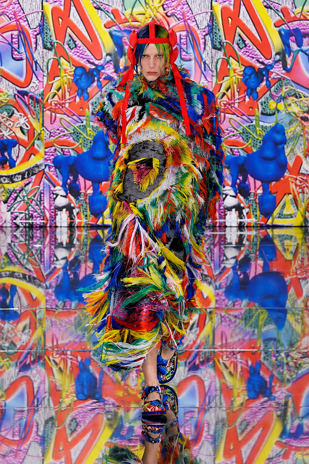 Look multicolor de Maison Margiela Artisanal 2019 de John Galliano con hilos, plumas y tocado rojo frente a un fondo de grafiti.