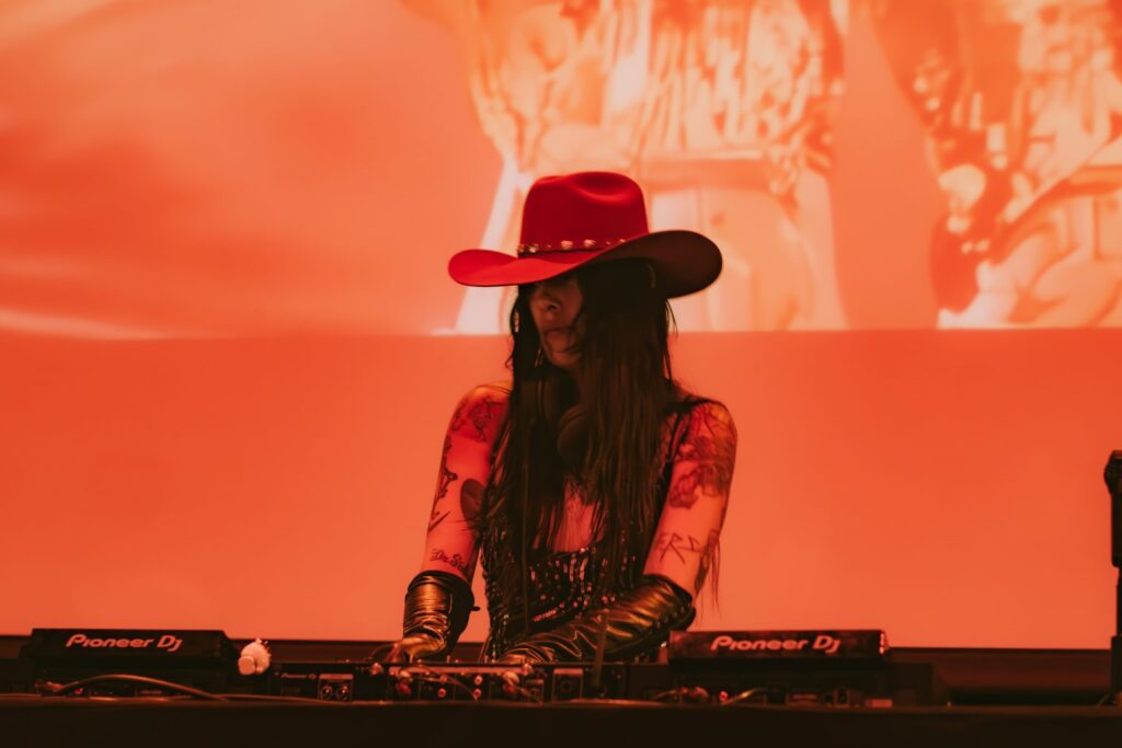 Rosa Pistola posa bajo luz roja con sombrero vaquero, una estética que conecta música de club y la nueva escena del tribal mexicano.