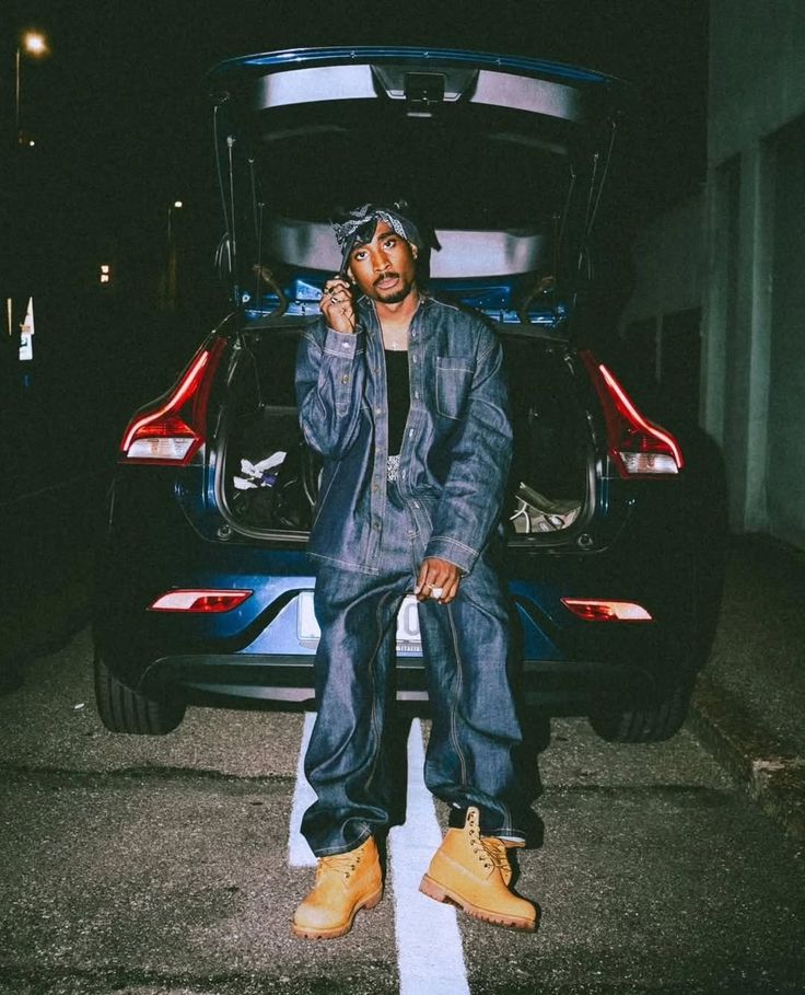 Tupac Shakur con conjunto holgado de mezclilla, bandana y botas de trabajo amarillas en una escena nocturna.