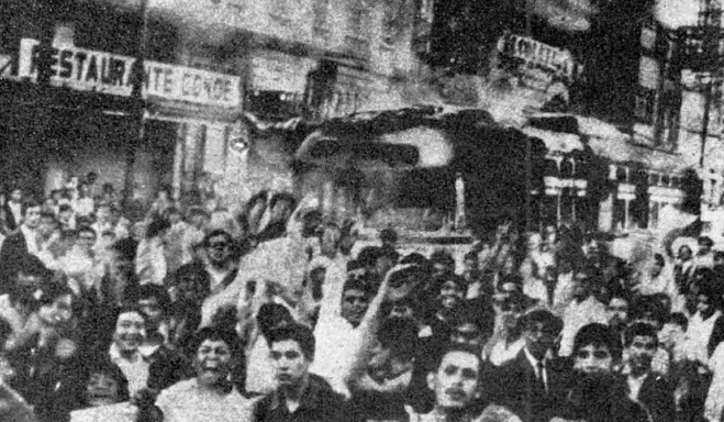 Multitud de jóvenes en una calle, varios con los brazos levantados y un autobús al fondo, en una fotografía en blanco y negro.