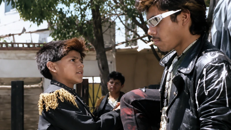 Dos jóvenes con estética punk frente a frente al aire libre, uno con chamarra de cuero y lentes blancos, en Corazones de humo.