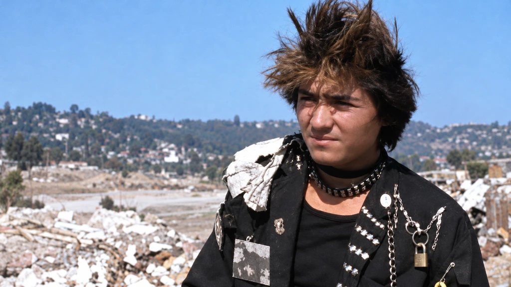 Joven punk con cabello levantado, estoperoles y cadenas, frente a escombros y viviendas en una ladera, en Corazones de humo.