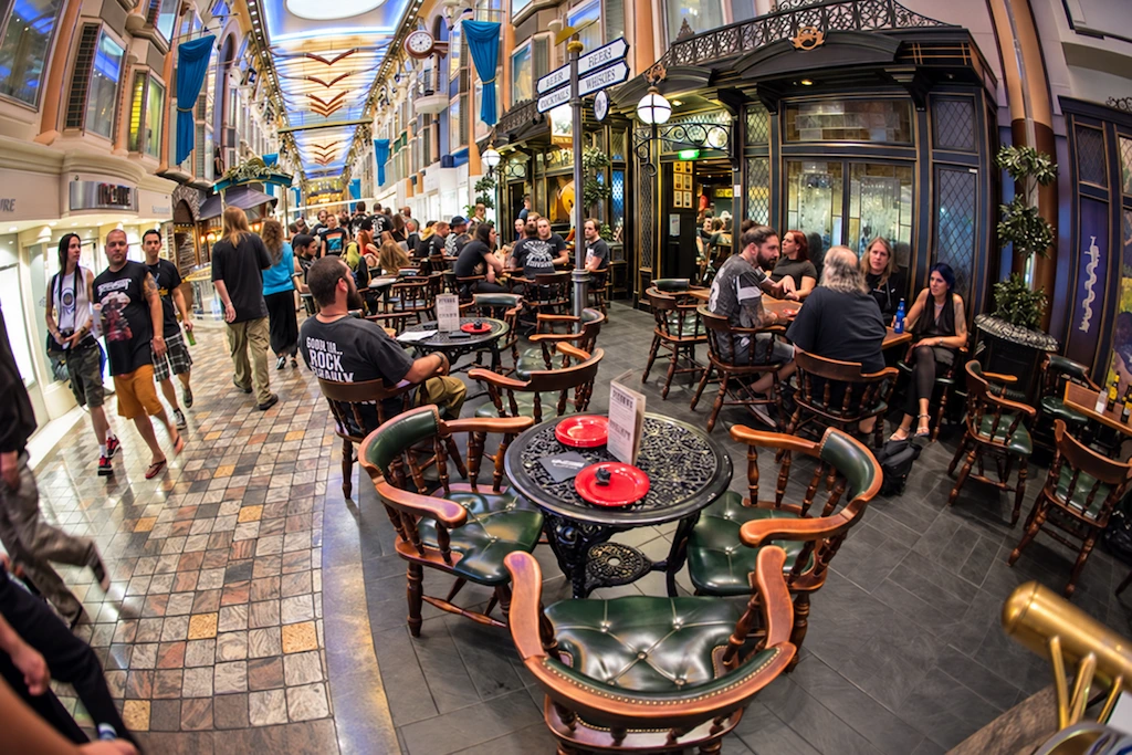 Pasajeros conversan en las mesas de un pub ubicado en una galería interior del crucero, entre comercios y fachadas decorativas.