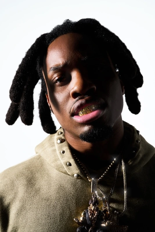 Denzel Curry en primer plano, con sudadera verde oliva, cadenas y dentadura dorada, sobre un fondo blanco.