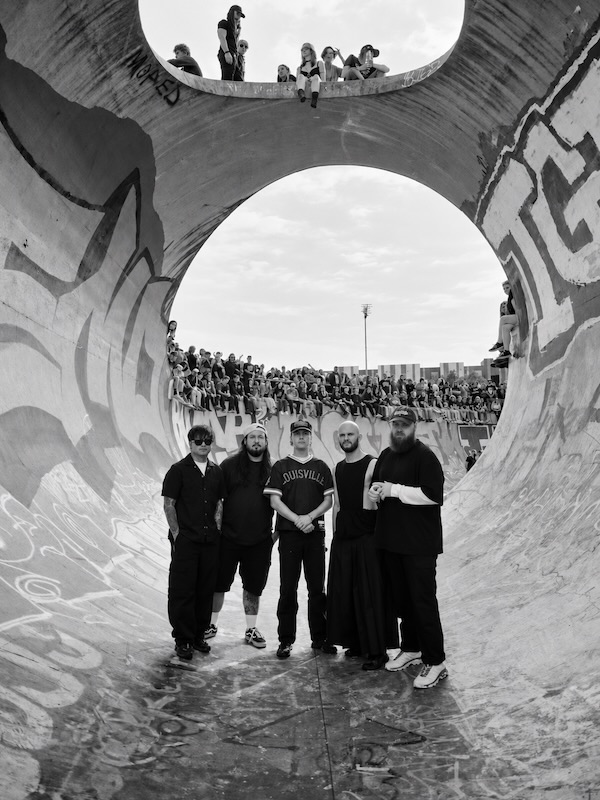 Los cinco integrantes de Knocked Loose posan en un skatepark con grafitis y público al fondo, en un retrato en blanco y negro.