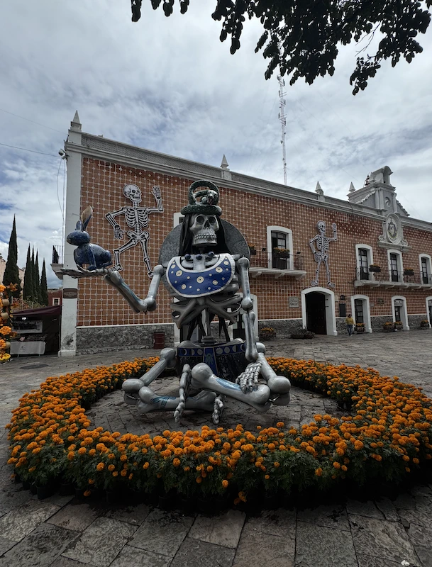 Figura esquelética monumental sentada, con adornos azules y una vasija, rodeada de cempasúchil en el Valle de Catrinas de Atlixco.